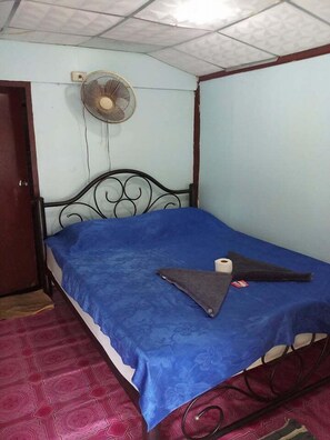 A single bed with a blue bedspread, a fan above it, and a patterned red carpet. - Royal Orchid Resort (Ko Pha-ngan)