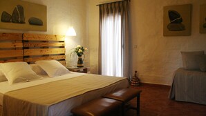 Une chambre d’hôtel avec une tête de lit en bois, un grand lit avec du linge de lit blanc, un banc au pied du lit, une table de chevet avec une lampe et une fenêtre avec des rideaux.