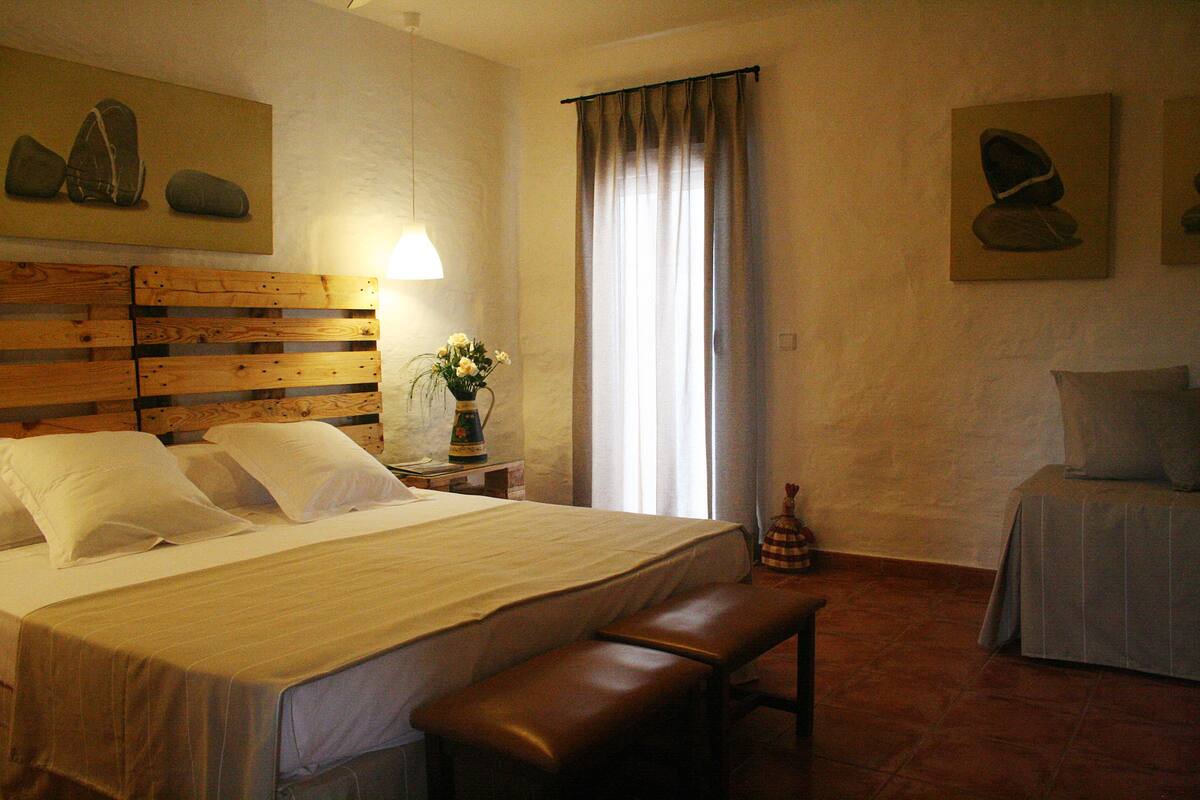 A hotel room with a wooden headboard, a large bed with white linens, a bench at the foot of the bed, a bedside table with a lamp, and a window with curtains.