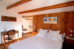Quarto com cabeceira de madeira, cama com roupa de cama branca, escrivaninha de madeira com cadeira e um quadro na parede.