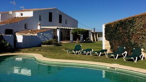 Un espace aménagé au bord de la piscine, avec des chaises longues, une piscine et un bâtiment blanc doté d’un toit en tuiles.