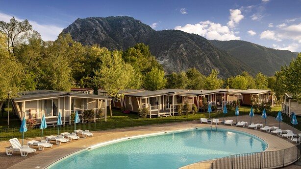 Un área de piscina con sillones de descanso y sombrillas, rodeada de edificios con montañas al fondo.