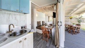Una cocina compacta con lavamanos y armarios, una zona de comedor con sillas de madera y una mesa, y una terraza exterior con pérgola.