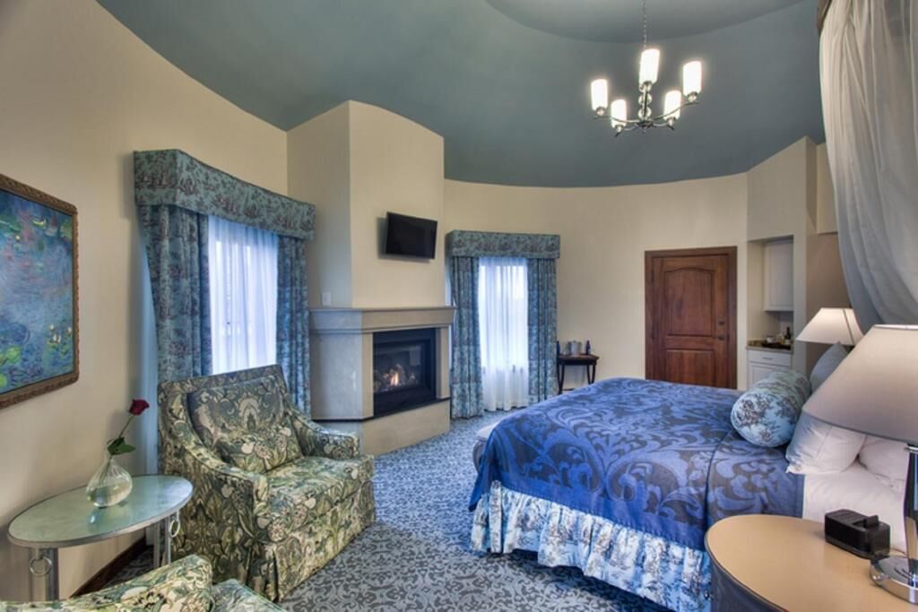 A bedroom with a fireplace, a bed with a blue patterned bedspread, a chair, a small table, and a television on the wall.