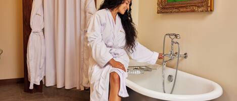 A person in a white bathrobe sitting by a bathtub in a bathroom with a painting on the wall.