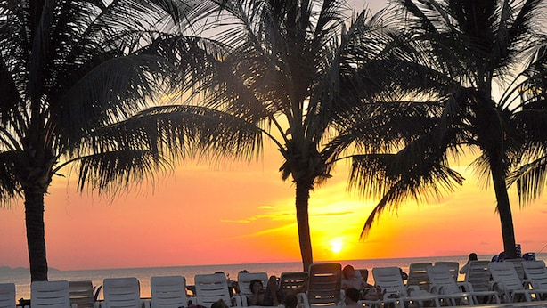 Un espace aménagé au bord de la piscine, avec des chaises longues et des palmiers, au moment du coucher du soleil.