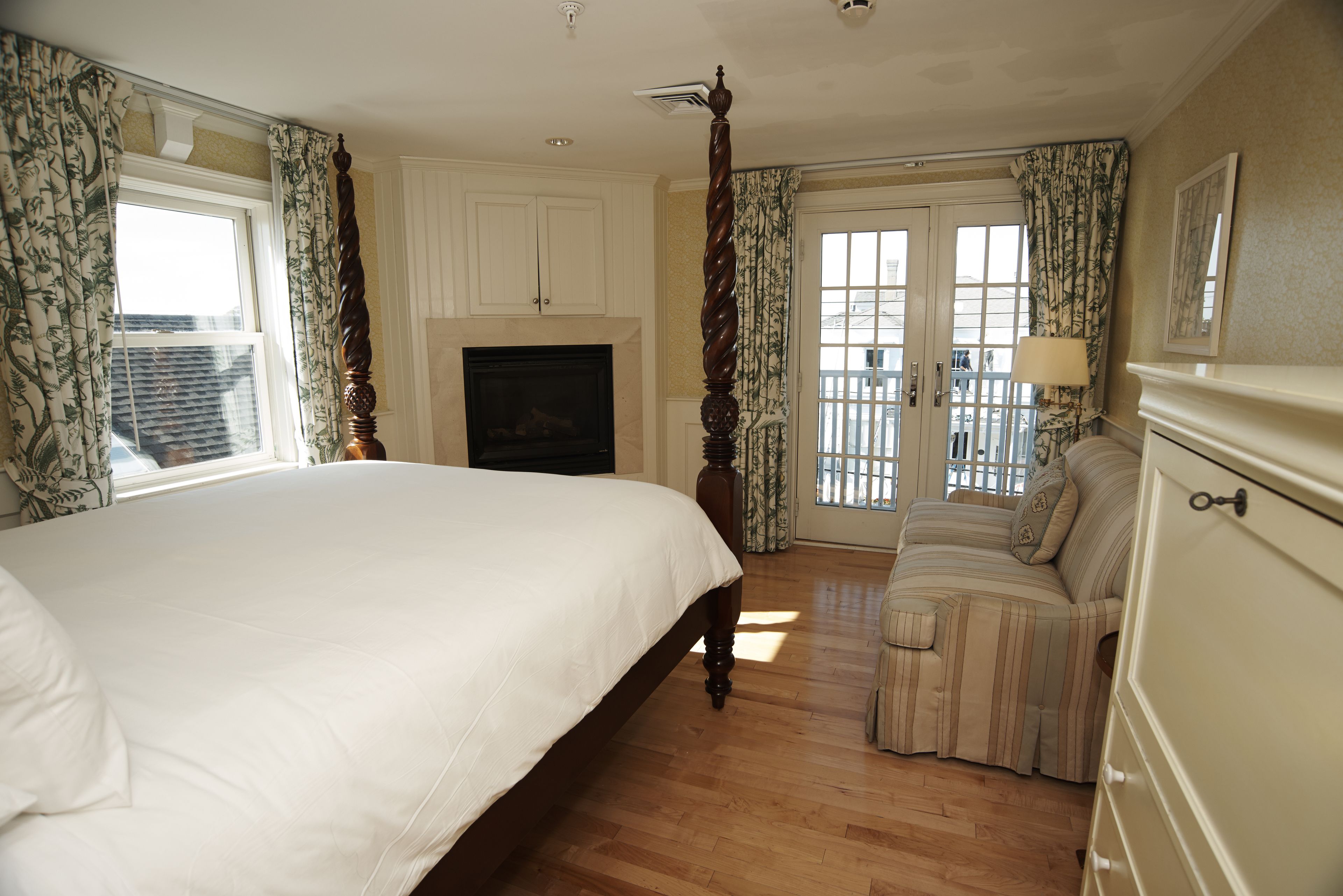 A bedroom with a large bed, a fireplace, a balcony with curtains, and a dresser.