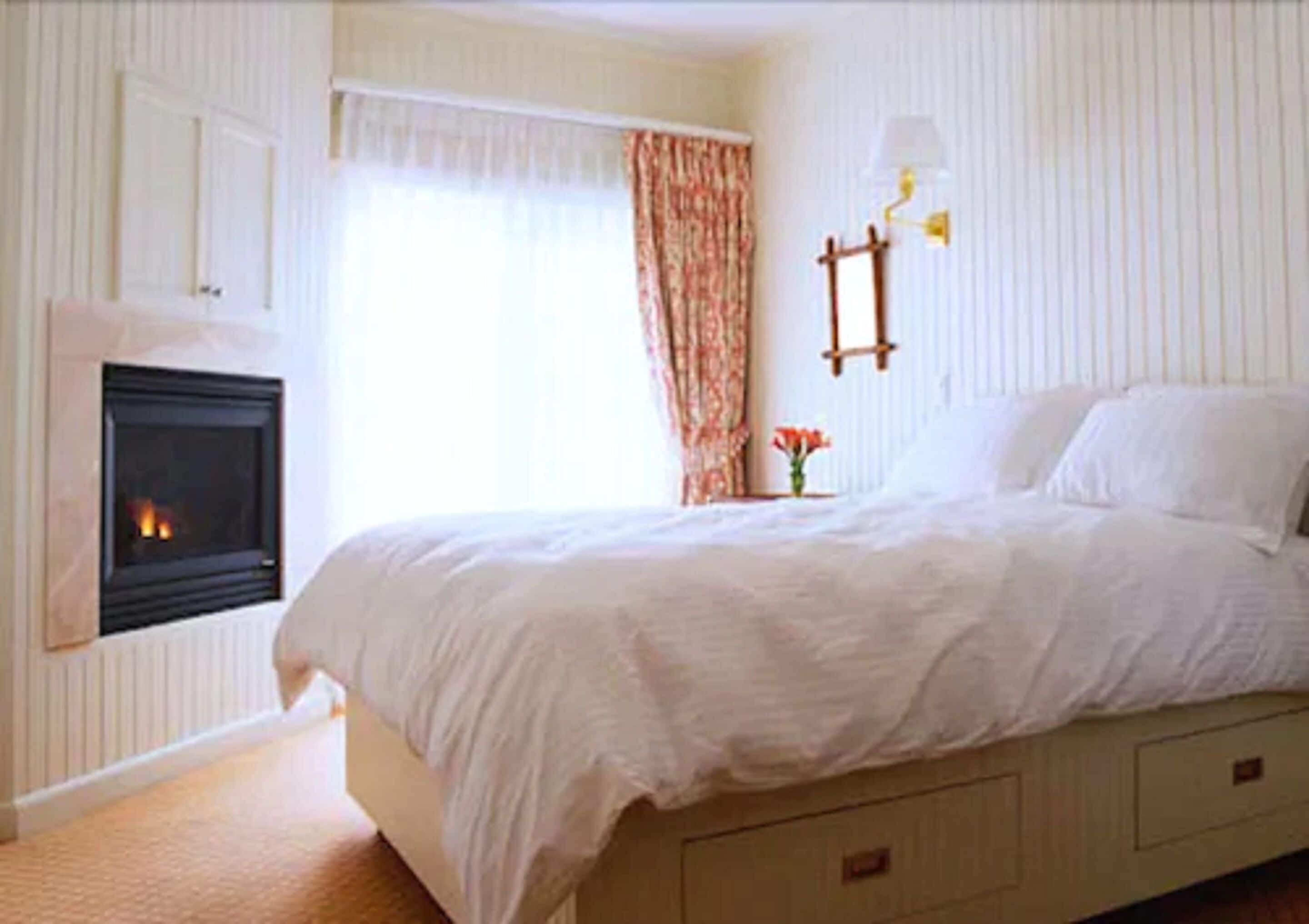 A bedroom with a bed, a fireplace, a window with curtains, and a wall-mounted lamp.