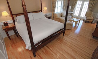 A four-poster bed with white linens, wooden nightstands with lamps, a sofa, and a small round table in a room with wooden flooring and large windows.