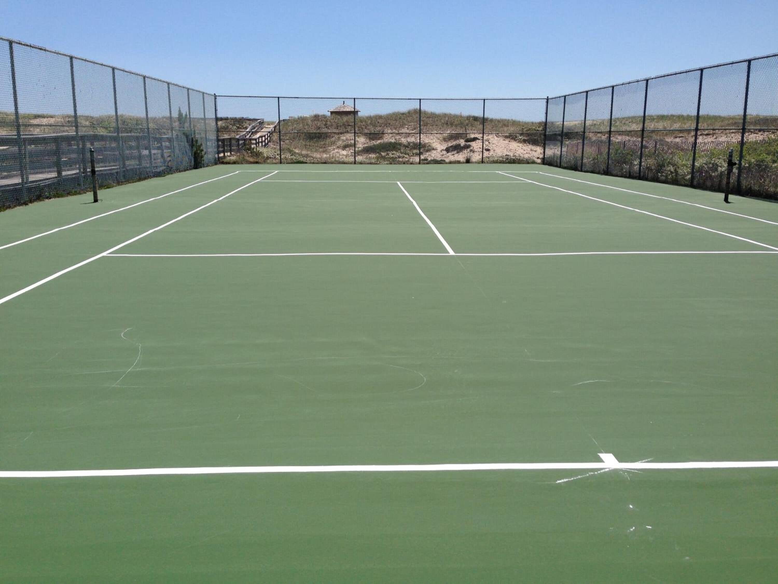A tennis court with a net, surrounded by a fence, and a sandy area in the background.
