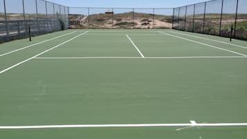A tennis court with a net, surrounded by a fence, and a sandy area in the background.