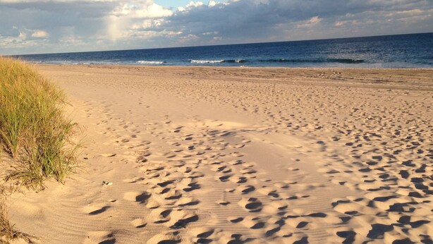 Ein Sandstrand mit Fußabdrücken, die zum Wasser führen, eine grasbewachsene Düne links und ein bewölkter Himmel.