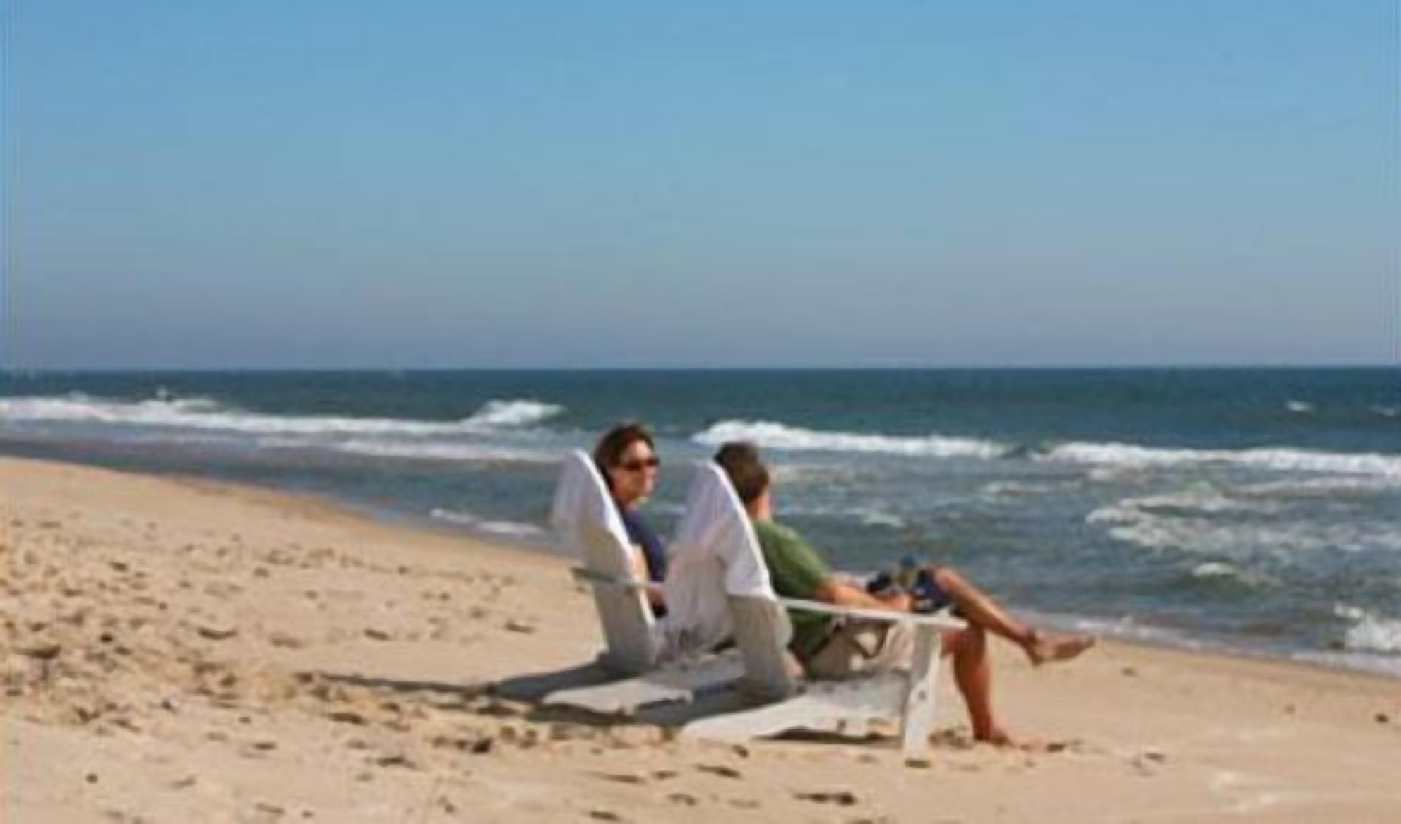 A beach scene with a person sitting on a beach chair and another person lying on the sand.