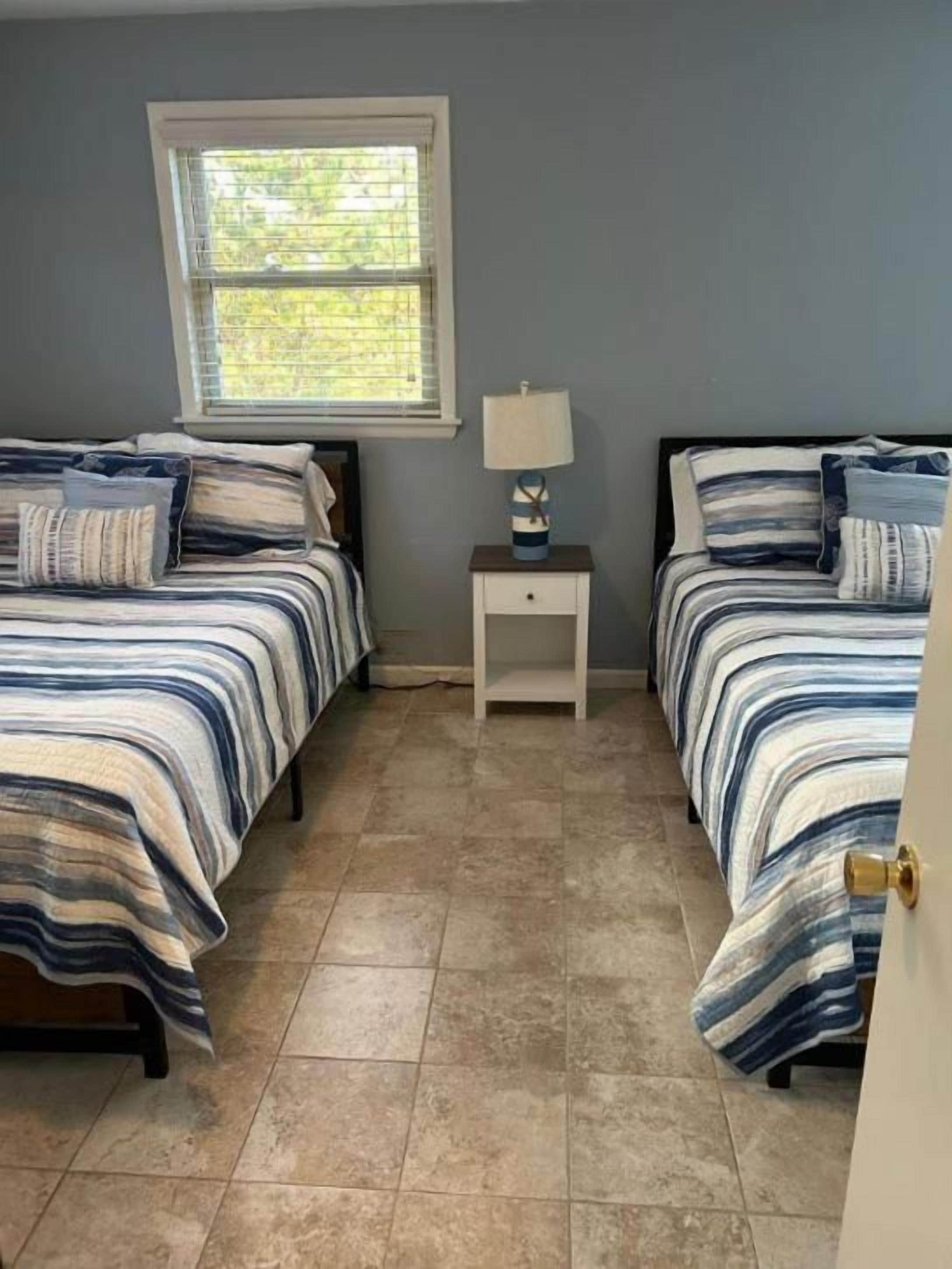 A bedroom with two beds, a nightstand with a lamp, and a window.