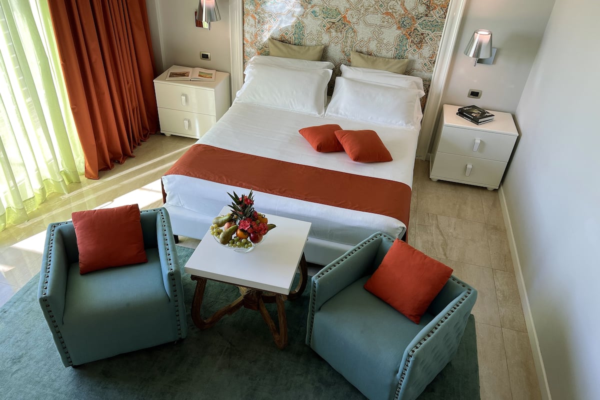 Une chambre d’hôtel avec un lit, deux tables de chevet, une petite table sur laquelle se trouve un panier de fruits, deux fauteuils et une tête de lit à motifs.