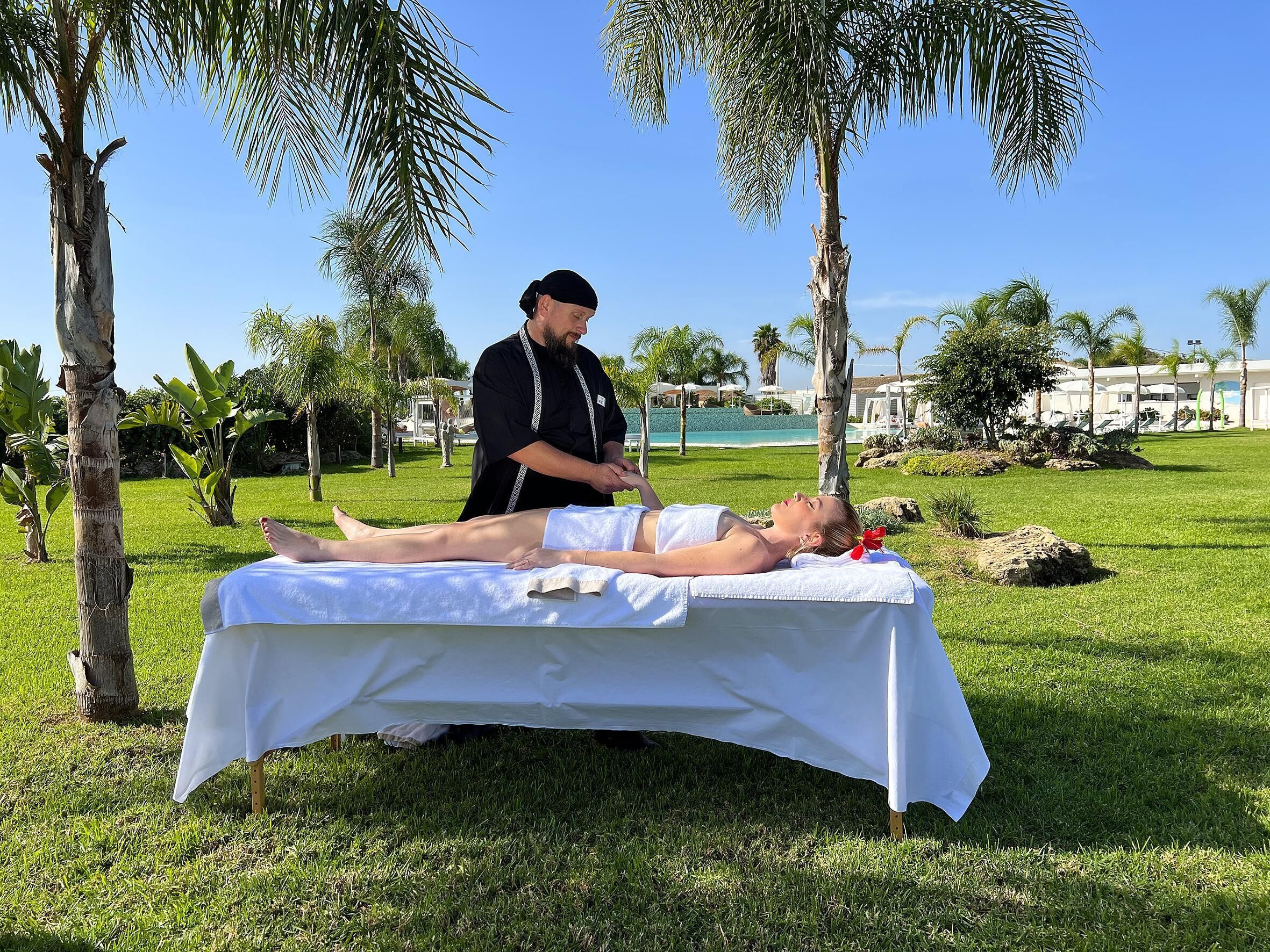 A massage therapy session taking place outdoors with a pool and palm trees in the background.