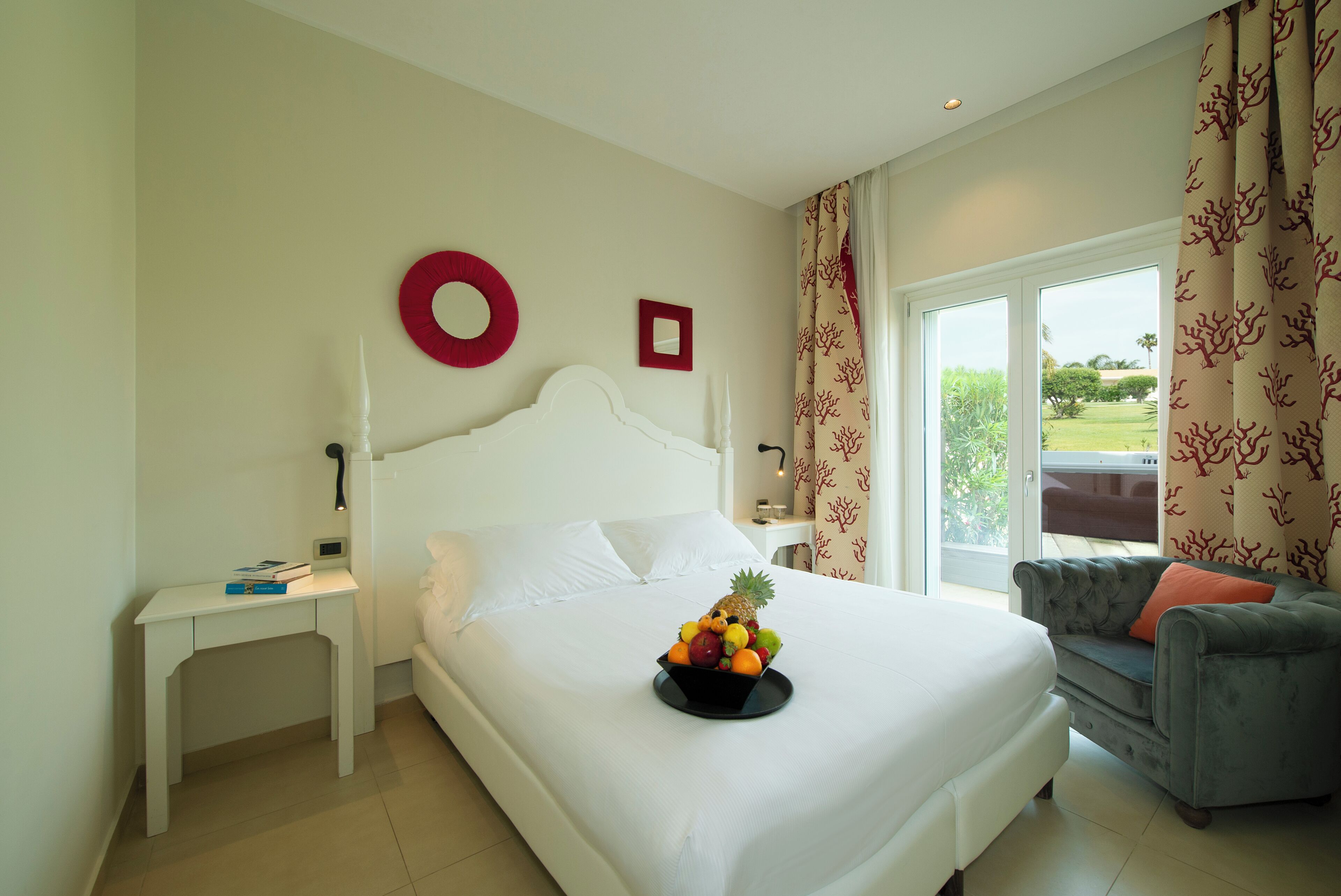 A hotel room with a bed, a bedside table, a fruit bowl, a grey sofa, and a view of the outdoors.