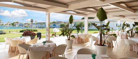 An outdoor dining area with tables and chairs, potted plants, and a view of a building and landscape.