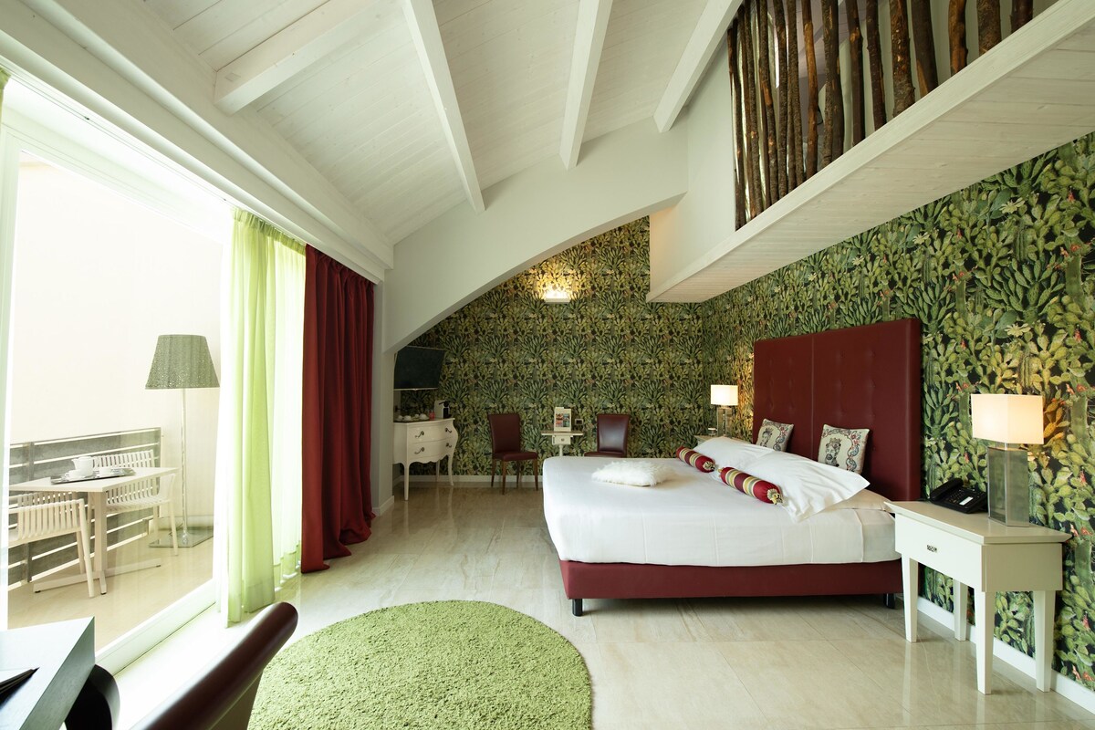 A bedroom with a large bed, a desk, and a chair. There is a window with curtains and a view of a balcony.