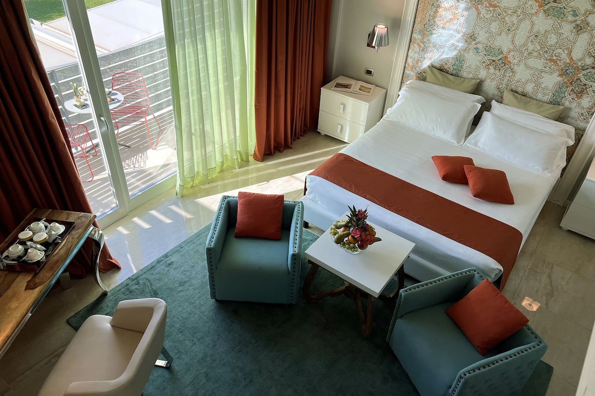 Une chambre d’hôtel avec un grand lit, deux fauteuils, une petite table sur laquelle se trouve un panier de fruits, une table de chevet et une fenêtre avec des rideaux.