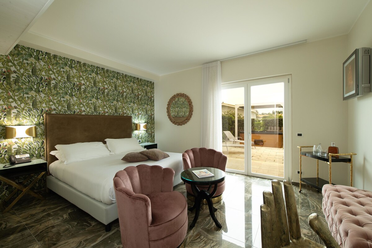 A hotel room with a bed, two pink chairs, a small table, a mirror, and a view of the outside.