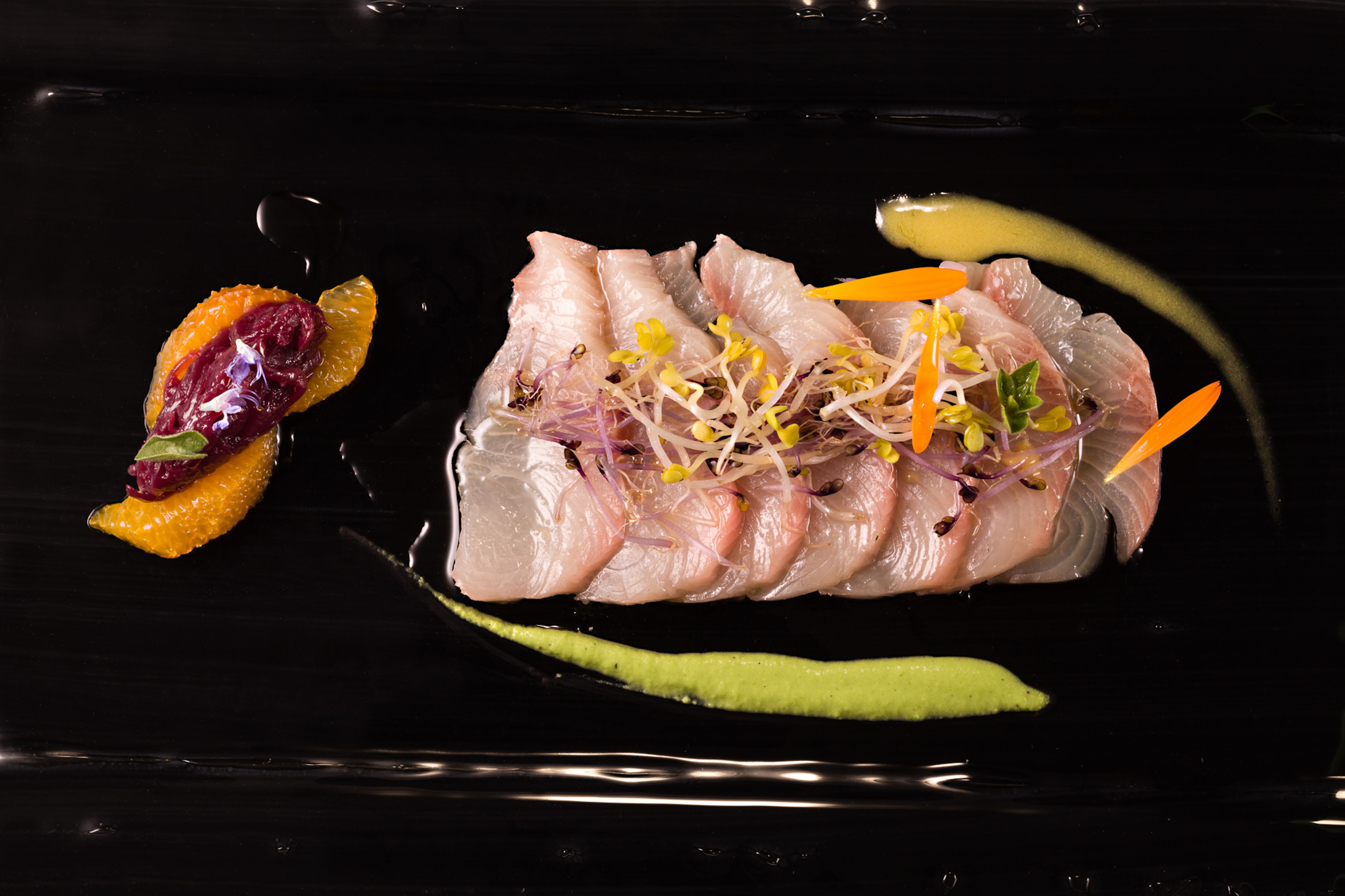 Ein schwarzer Teller mit rohem Fisch, Orangenscheiben und Sprossen.