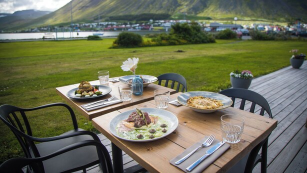 An outdoor dining table with plates of food, glasses, and chairs on a wooden deck with a view of a mountain and a body of water in the background. - Hótel Ísafjörður (Isafjordur)
