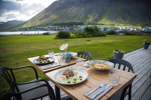Mesa al aire libre con platos de comida, vasos y sillas sobre una terraza de madera, con vista a una montaña y un cuerpo de agua al fondo.