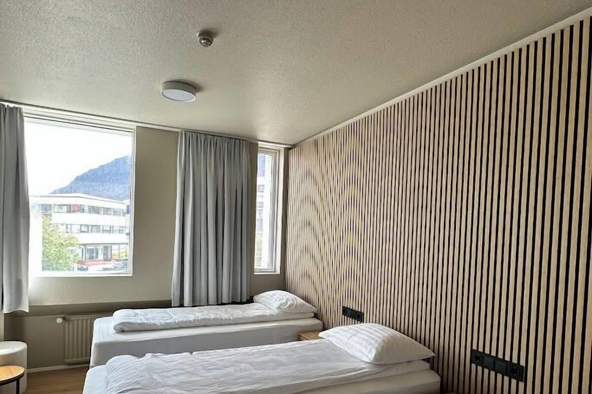 Ein Hotelzimmer mit drei Betten, einem Vorhangfenster, einem Heizkörper und Blick auf ein Gebäude.