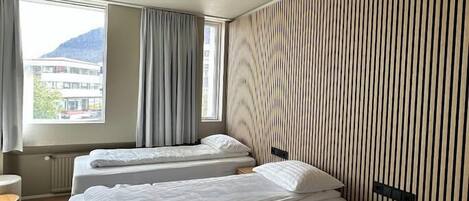 A hotel room with three beds, a window with curtains, a radiator, and a view of a building outside.