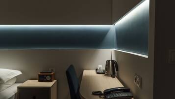A hotel room with a bed, a bedside table with a radio, a desk with a telephone, a cup, and a kettle.
