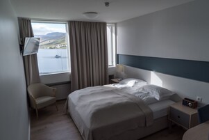 Habitación de hotel con cama, una silla, mesita de noche, lámpara y vistas al mar.