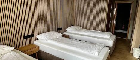 A hotel room with three beds, a wooden slatted wall, a small wooden nightstand, and a ceiling light.
