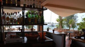 Un espace bar avec des étagères remplies de bouteilles d’alcool, un comptoir et des places assises en plein air offrant une vue sur la mer et les bateaux.