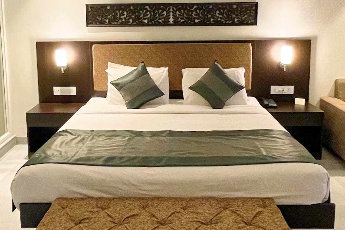 Une chambre d’hôtel avec un grand lit, deux tables de chevet, une petite table, un banc et un tapis rouge à motifs.