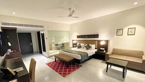 A modern hotel room with a bed, a desk, a chair, a sofa, a glass-enclosed shower, and a ceiling fan.