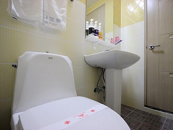 A bathroom with a toilet, sink, and shower. There are toiletries on the shelf above the sink. - Brown Hotel (Suncheon)