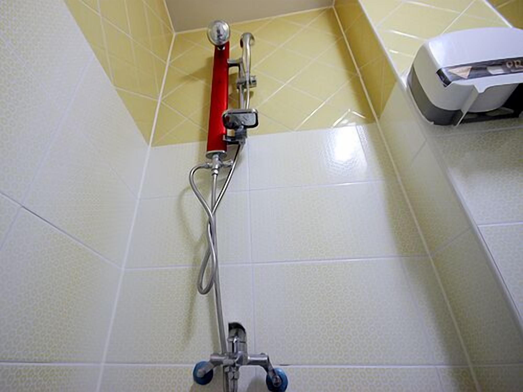 A shower with a red handle, a white wall, and yellow-tiled floor.