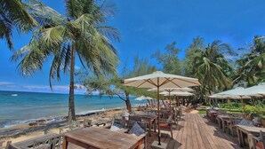 Un restaurante en la playa con mesas y sillas de madera, sombrillas y palmeras.