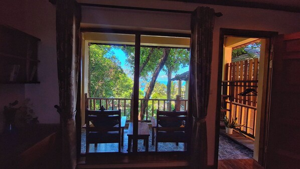 A room with a large sliding glass door leading to an outdoor area with a view of trees and a building. There are wooden benches and a table inside the room. - Ancarine Beach Resort (Phu Quoc)