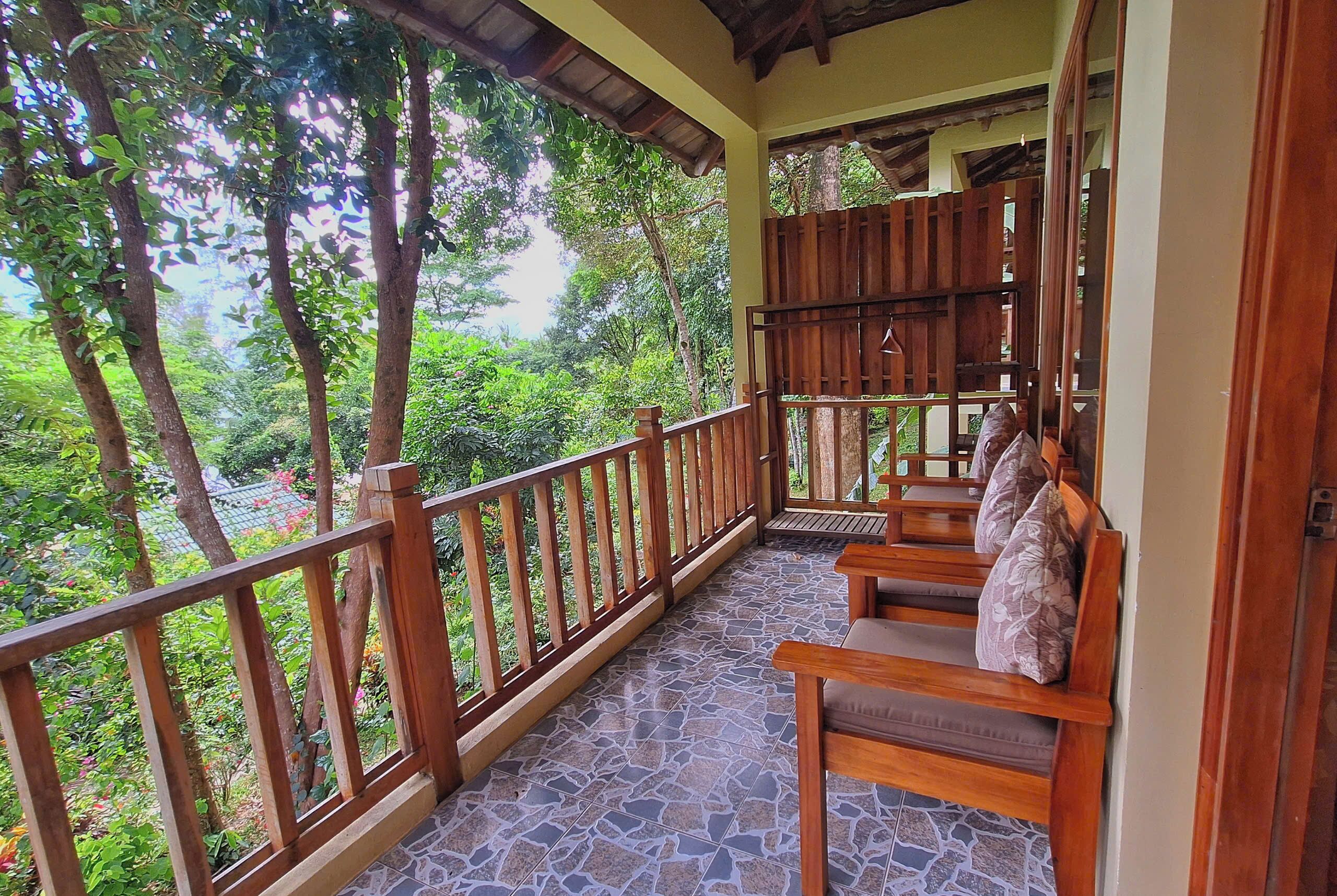 A wooden balcony with a tiled floor, wooden railings, and a seating area with cushions.