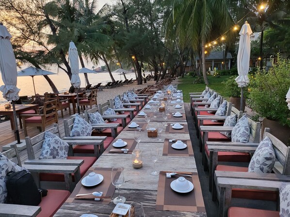Un restaurante frente al mar con mesas y bancos de madera, vajilla blanca y luces colgantes.
