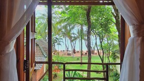 Vista a través de una ventana con cortinas blancas, que muestra un balcón de madera y un jardín exuberante con árboles y un tejado a la distancia.