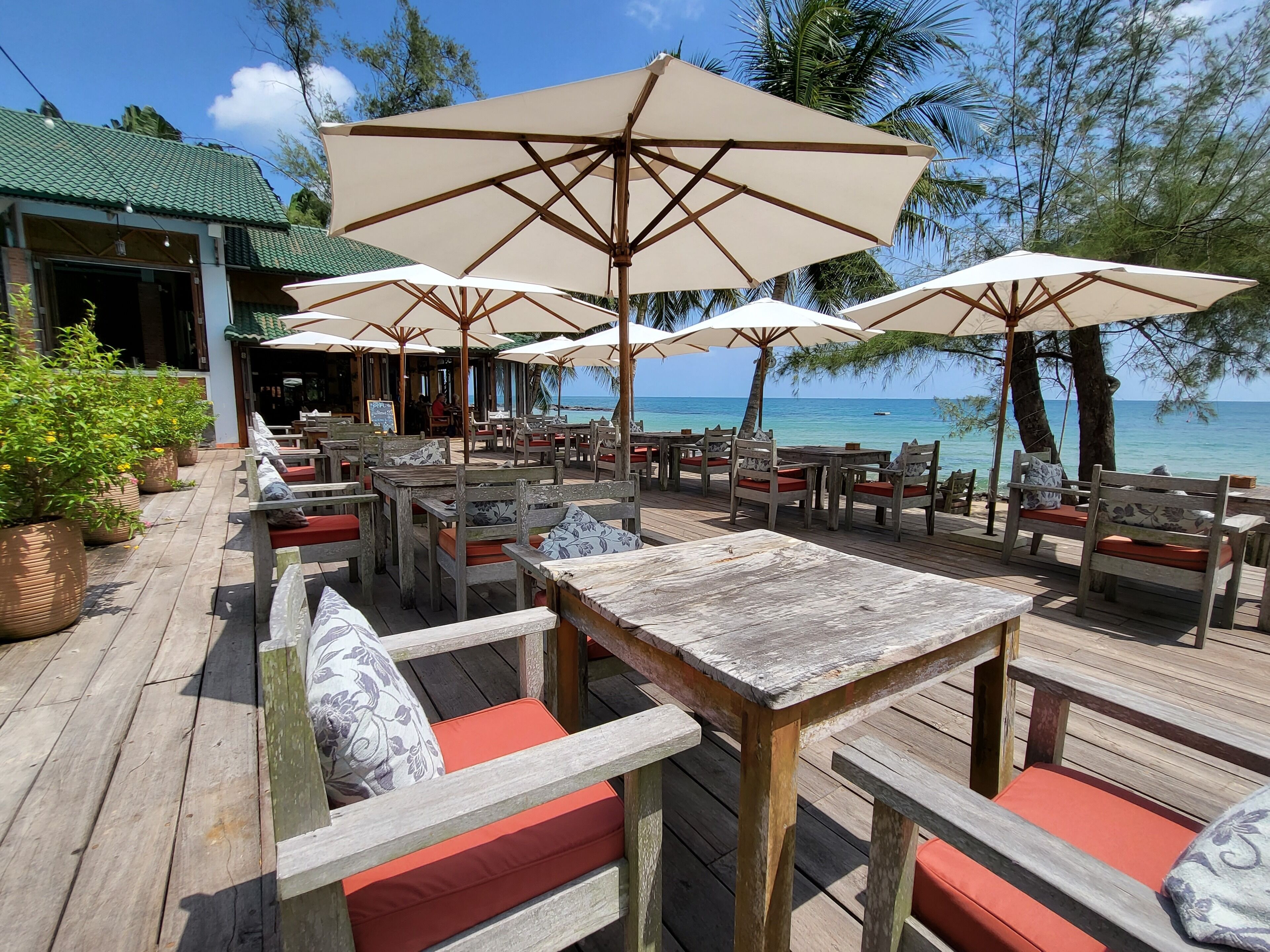 An outdoor seating area with wooden tables and chairs, large umbrellas, and a view of the ocean.