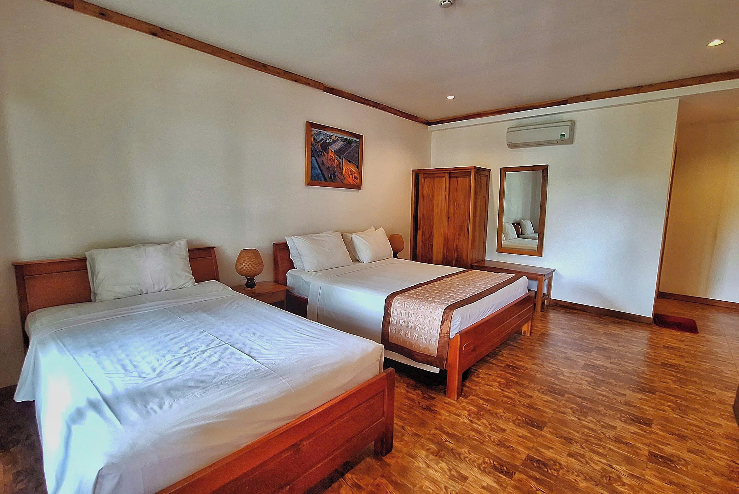 Habitación de hotel con dos camas de madera, un armario de madera, una mesita de noche con lámpara y un aire acondicionado de pared.
