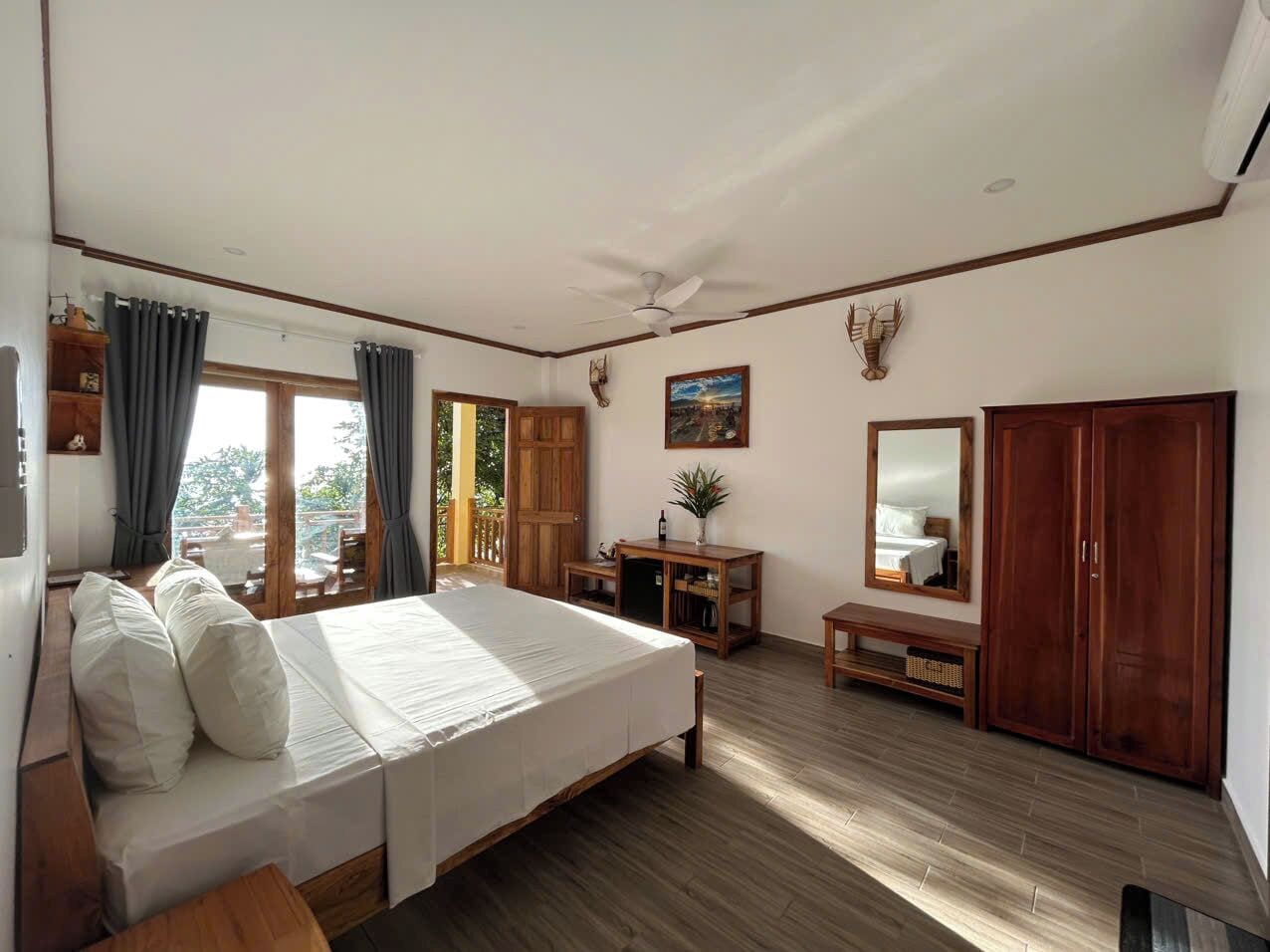 A spacious bedroom with a large bed, wooden flooring, a ceiling fan, a wooden wardrobe, a small table with a plant, and a window with curtains.