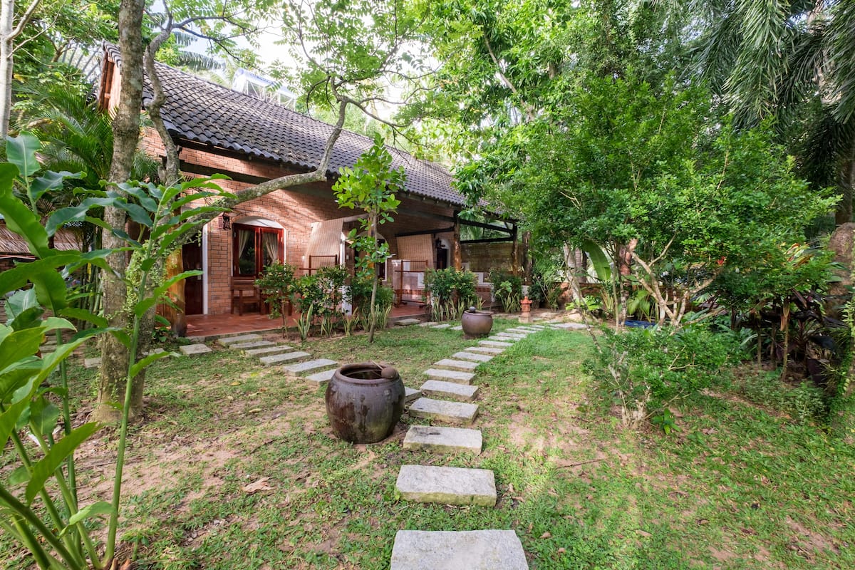 A rustic house with a tiled roof, surrounded by lush greenery and a stone pathway leading to the entrance.