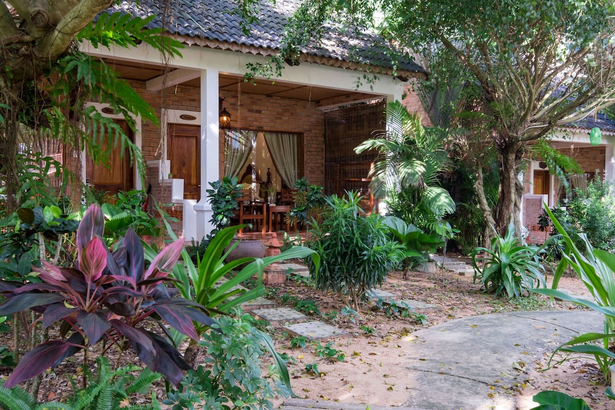 A traditional house with a covered porch, surrounded by lush greenery and a stone pathway.