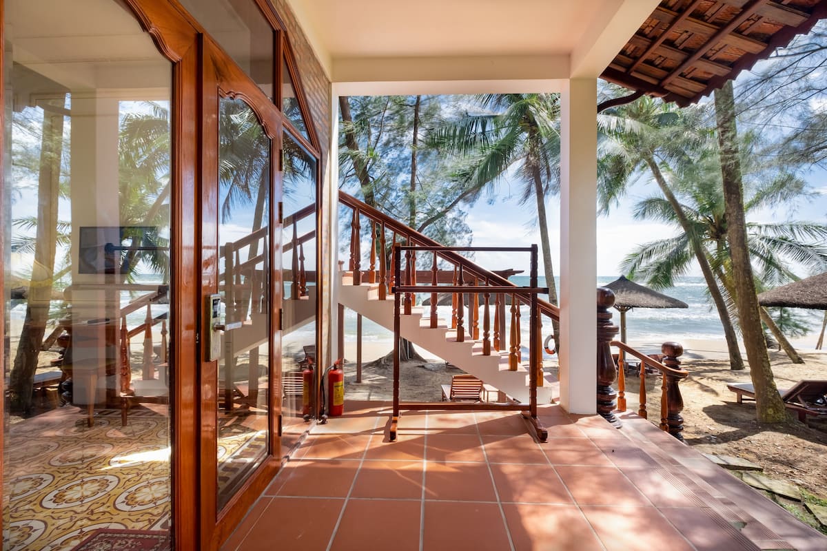 A beachside hotel room with a large glass door, a wooden staircase, and a view of the beach and palm trees.