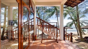 A beachside hotel room with a large glass door, a wooden staircase, and a view of the beach and palm trees. - Coco Palm Beach Resort & Spa (Phu Quoc)